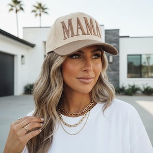 May include: A beige baseball cap with a brown brim and the word "MAMA" embroidered in brown. The cap is worn by a person wearing a white t-shirt and gold necklaces.