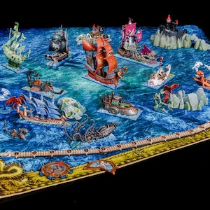 Puede incluir: Colección de barcos piratas en miniatura y criaturas marinas sobre un tapete de juego azul con temática oceánica. Los barcos son de varios colores, incluyendo rojo, negro y verde. El tapete tiene un borde detallado con elementos náuticos.