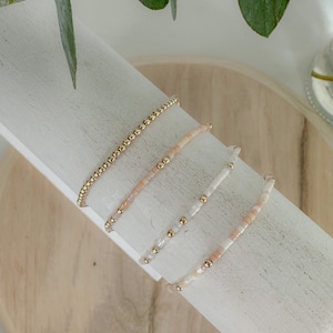 May include: Four bracelets stacked on a white display stand. The top bracelet is gold, the next is peach, the next is white with gold accents, and the bottom bracelet is peach and white.