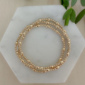 May include: Three gold beaded bracelets stacked on a white marble surface. The beads are oval shaped and have a smooth, polished finish.