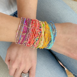 May include: A collection of colorful beaded bracelets in various shades, including pink, orange, yellow, and blue. Each bracelet features small, colorful beads and gold-colored accents. The bracelets are stacked on a wrist, creating a vibrant and eye-catching display.