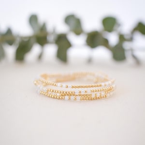 May include: Stack of beaded bracelets. The bracelets feature a combination of small, gold-colored beads and white beads. The bracelets are stacked on top of each other, creating a layered effect. The background is blurred, with green foliage.