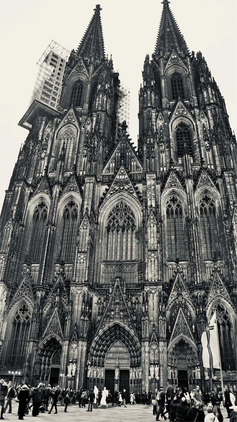 K&ouml;nnte beinhalten: Ein Schwarzwei&szlig;foto des K&ouml;lner Doms in Deutschland. Der Dom ist ein gro&szlig;es gotisches Bauwerk mit zwei hohen T&uuml;rmen. Das Foto wurde vom Boden aus aufgenommen und zeigt den Dom von unten. Im Vordergrund des Fotos sind Menschen zu sehen.