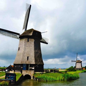 May include: Two traditional Dutch windmills with wooden blades stand in a field with a canal running through it. The sky is cloudy and the windmills are in the foreground.