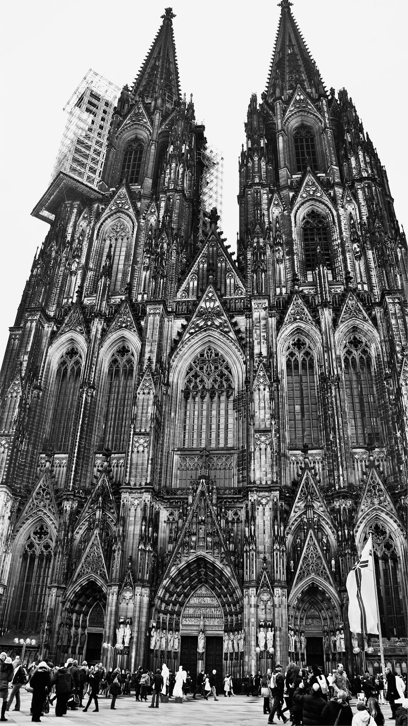 K&ouml;nnte beinhalten: Ein Schwarzwei&szlig;foto des K&ouml;lner Doms in Deutschland. Der Dom ist ein gro&szlig;es gotisches Bauwerk mit zwei hohen T&uuml;rmen. Die Vorderseite des Doms ist mit aufwendigen Schnitzereien und Buntglasfenstern verziert. Vor dem Dom gehen Menschen spazieren.