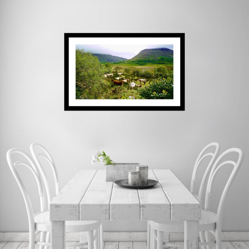 May include: A framed landscape print featuring a scenic view of a valley with grazing cattle. The artwork showcases green hills, a cloudy sky, and lush vegetation. The print is displayed on a white table with white chairs.