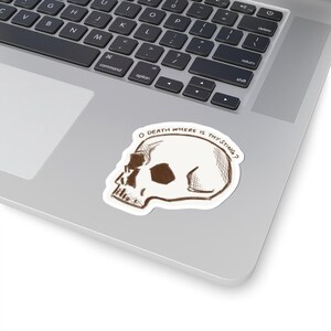 以下が含まれることがあります： 白いステッカーに、茶色の線で描かれた頭蓋骨と「O DEATH WHERE IS THY STING?」というテキストが書かれています