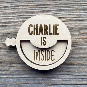 May include: A round, light-colored wooden sign with the engraved text "CHARLIE IS INSIDE". The sign has a semi-circular cutout and a small handle. The background is a weathered wooden surface.