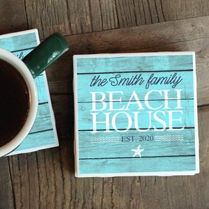 May include: Two square coasters with a teal wood grain design and white text that reads "the Smith family BEACH HOUSE EST. 2020". A white mug with a green handle sits on one coaster, filled with dark liquid.