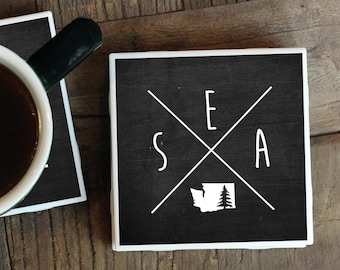 May include: Two black and white coasters with a white outline. One coaster has a white mug with black coffee on it. The other coaster has a white outline of the state of Washington with a tree inside and the letters "SEA" with a white X in the middle.