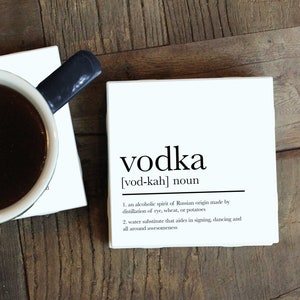 May include: A white coaster with the word "vodka" printed in black, with the pronunciation and definition of the word below. The coaster is on a wooden surface with a cup of coffee on a coaster to the left.