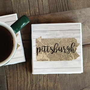 May include: Two white ceramic coasters featuring a vintage map design of Pittsburgh, Pennsylvania. The coasters have a wood grain background and the word "Pittsburgh" in black script.