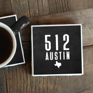 May include: Two black square coasters with white text and a white outline. The top coaster says "512 Austin" with a white outline of the state of Texas. The bottom coaster has a white coffee mug with black coffee on top.