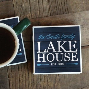 May include: Two square coasters with a blue and white design. The top coaster says "the Smith family LAKE HOUSE EST. 2018" in white text with blue accents. The bottom coaster is white with a green mug on top. The mug is filled with dark coffee.