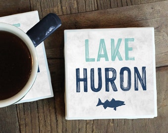 Puede incluir: Dos posavasos de cerámica blanca con texto azul y verde. El posavasos de la izquierda tiene una taza de café encima. El posavasos de la derecha dice "LAKE HURON" con una silueta de pez azul.