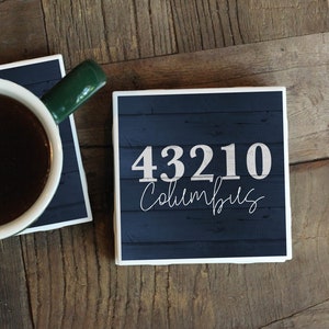 May include: Two square coasters with a dark blue woodgrain design. One coaster has a cup of coffee on it. The other coaster has the text "43210 Columbus" in white.