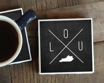Puede incluir: Dos posavasos de cerámica en blanco y negro con un contorno blanco. El posavasos de la derecha tiene un contorno blanco del estado de Kentucky con las letras "LOU" en forma de cruz encima. El posavasos de la izquierda tiene una taza de café encima.