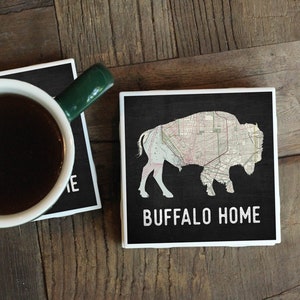 May include: Two black and white coasters with a white border. One coaster features a silhouette of a buffalo with a map pattern inside. The other coaster has the text "BUFFALO HOME".