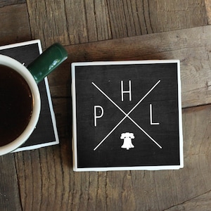 May include: Two black and white coasters with a white graphic of a bell and the letters "H", "P", "L" on a black background. The coasters are on a wooden surface with a cup of coffee on one of the coasters.