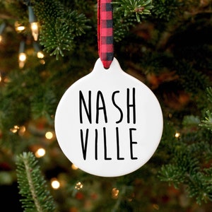 May include: White ceramic Christmas ornament with black text that reads "NASHVILLE". The ornament is hanging from a red and black buffalo plaid ribbon on a green Christmas tree.