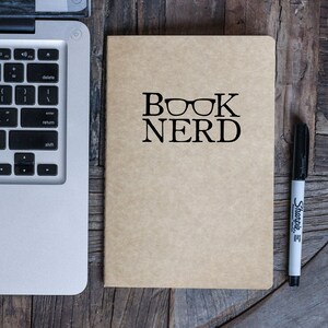 May include: A brown notebook with the text "BOOK NERD" and a pair of glasses printed on the cover. A black Sharpie marker is lying on a wooden surface next to the notebook.