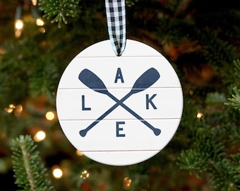 Puede incluir: Adorno navideño redondo blanco con una cinta azul y blanca a rayas. El adorno tiene un fondo de madera blanca con una silueta azul de dos remos cruzados y la palabra "LAKE" en letras azules.