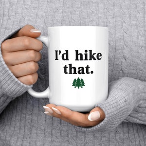 May include: White ceramic mug with black text that reads "I'd hike that." and a green silhouette of three pine trees.