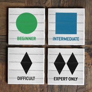 May include: Four white square coasters with black text and different colored shapes. The first coaster has a green circle and the text "BEGINNER". The second coaster has a blue square and the text "INTERMEDIATE". The third coaster has a black diamond and the text "DIFFICULT". The fourth coaster has two black diamonds and the text "EXPERT ONLY".