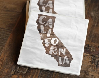 Op de afbeelding: Twee witte keramische onderzetters met een houtnerfdesign met de omtrek van de staat Californië en het woord "CALIFORNIA" in witte letters.