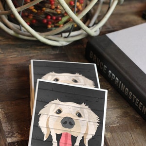 Puede incluir: Dos posavasos cuadrados de cer&aacute;mica con fondo de madera negra y contorno blanco. El posavasos superior presenta un perro golden retriever con la lengua fuera. El posavasos inferior es parcialmente visible.