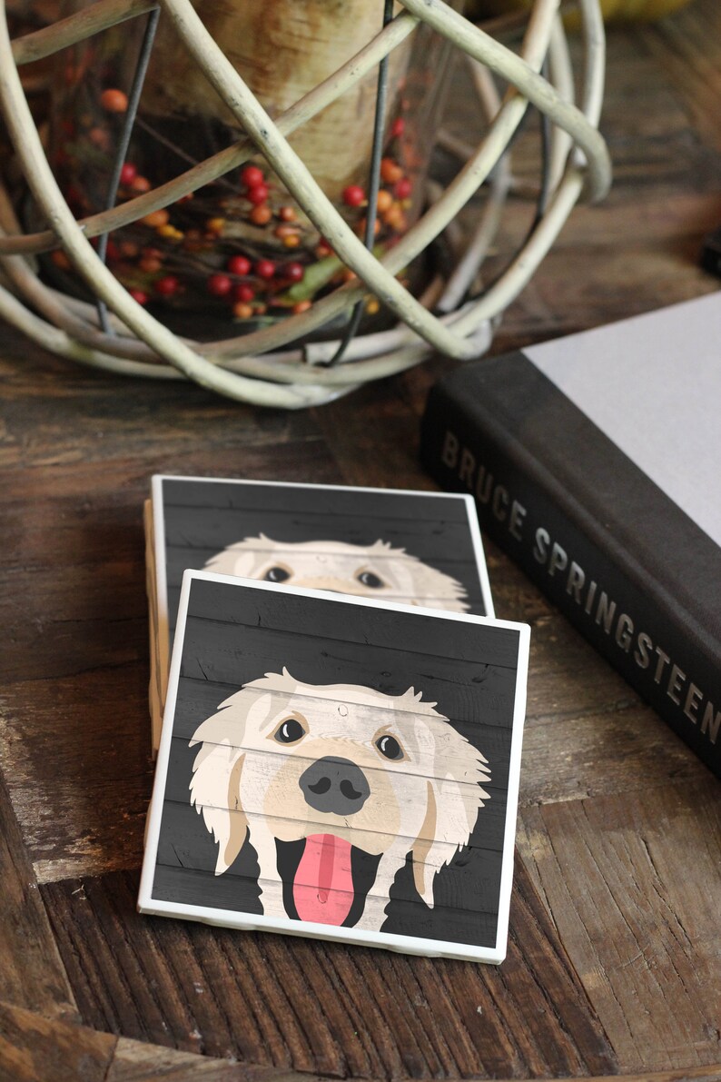 Puede incluir: Dos posavasos cuadrados de cer&aacute;mica con fondo de madera negra y contorno blanco. El posavasos superior presenta un perro golden retriever con la lengua fuera. El posavasos inferior es parcialmente visible.