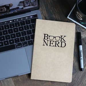 May include: A brown notebook with the text "BOOK NERD" and a pair of glasses printed on the front. A black marker and a laptop are also in the image.