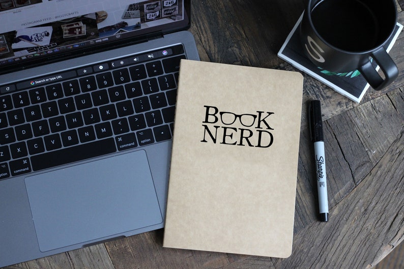May include: A brown notebook with the text "BOOK NERD" and a pair of glasses printed on the front. A black marker and a laptop are also in the image.