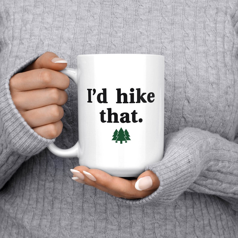 May include: White ceramic mug with black text that reads "I'd hike that." and a green silhouette of three pine trees.