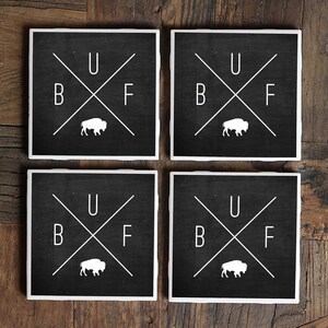 May include: Set of four black square coasters with white lettering and a white buffalo silhouette. The coasters read "B U F F" with the letters arranged in a cross pattern.