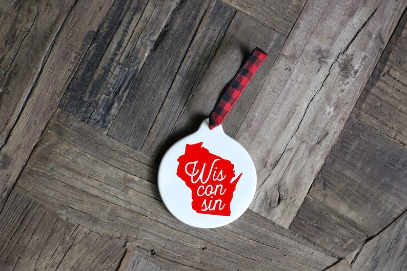 May include: White ceramic ornament shaped like the state of Wisconsin with the words "Wiscon sin" written in red inside the outline of the state. The ornament has a red and black plaid ribbon attached.