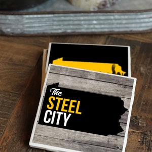 May include: Two black and white coasters with a wood grain design. The top coaster features a yellow outline of the state of Pennsylvania. The bottom coaster features the text "The Steel City" in yellow letters.