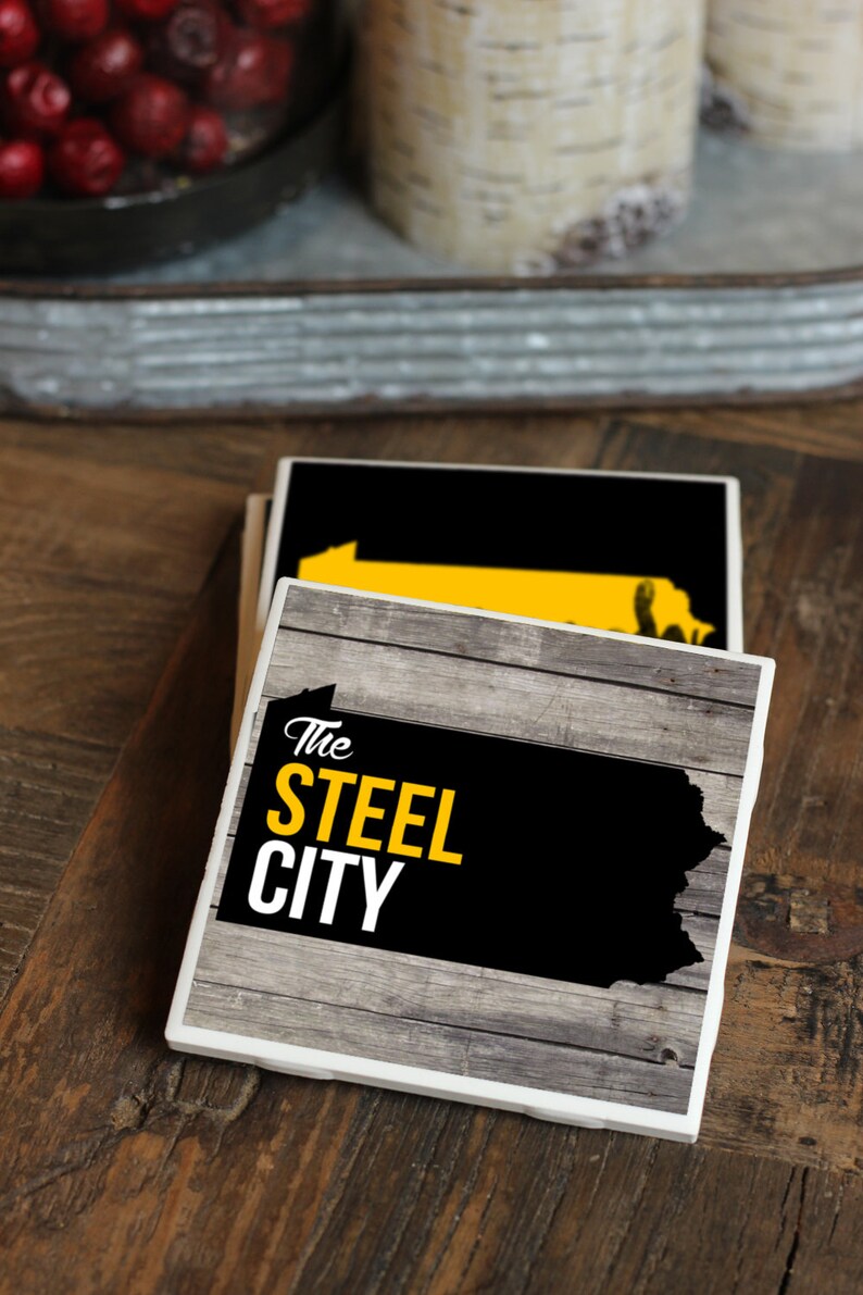 May include: Two black and white coasters with a wood grain design. The top coaster features a yellow outline of the state of Pennsylvania. The bottom coaster features the text "The Steel City" in yellow letters.