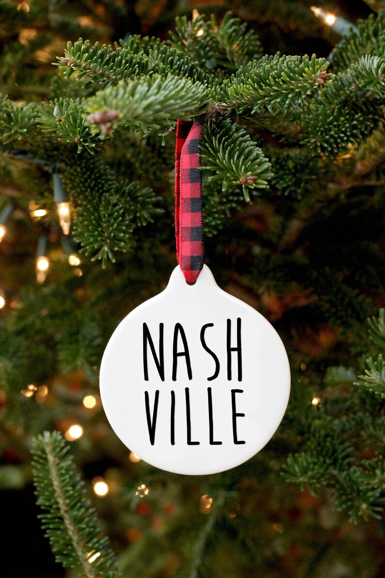 May include: White ceramic Christmas ornament with black text that reads "NASHVILLE". The ornament is hanging from a red and black buffalo plaid ribbon on a green Christmas tree.