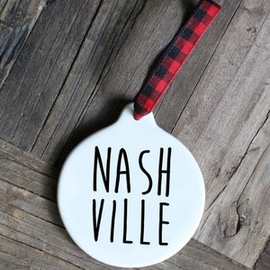 May include: White ceramic ornament with black text that reads "NASHVILLE". The ornament has a red and black buffalo plaid ribbon attached.