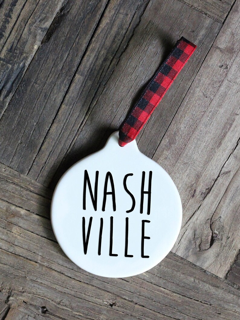 May include: White ceramic ornament with black text that reads "NASHVILLE". The ornament has a red and black buffalo plaid ribbon attached.
