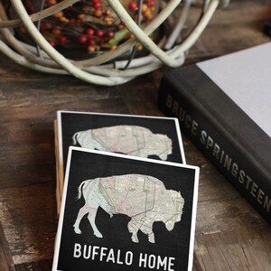 May include: Two black coasters with a white outline. The top coaster features a map of Buffalo, New York, in the shape of a buffalo. The bottom coaster has the text "BUFFALO HOME" in white.
