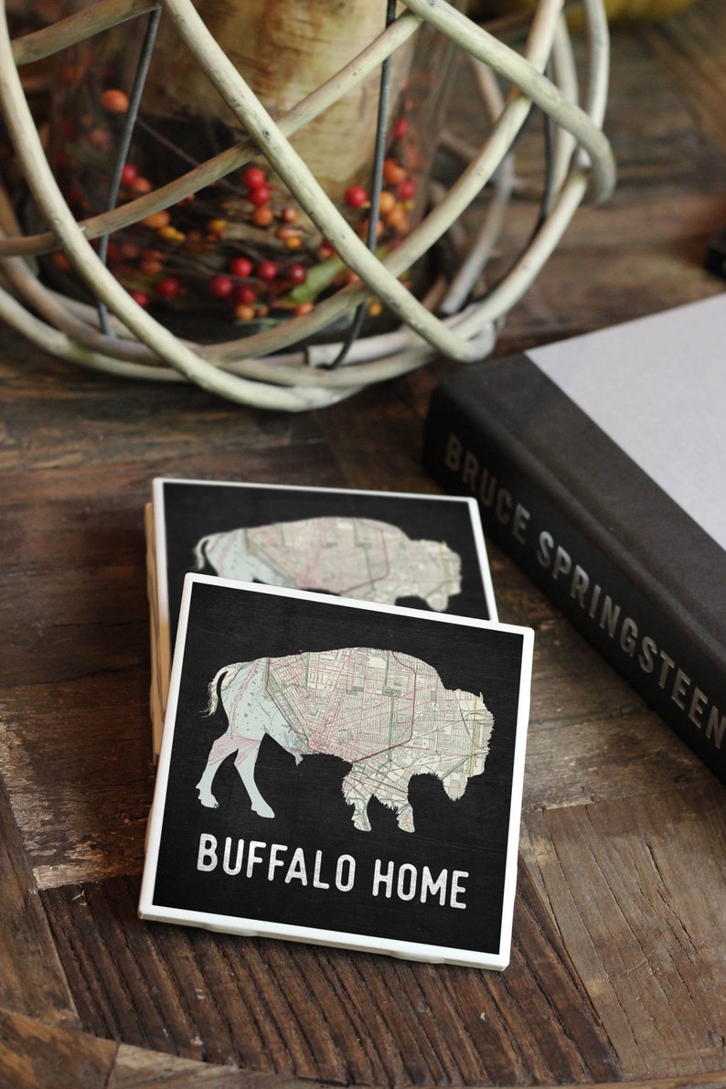 May include: Two black coasters with a white outline. The top coaster features a map of Buffalo, New York, in the shape of a buffalo. The bottom coaster has the text "BUFFALO HOME" in white.