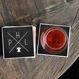 May include: Two black square coasters with white lettering. The coasters feature the letters "H", "P", "L" and a stylized Liberty Bell. One coaster has a glass of red liquid on top of it.