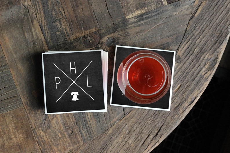 May include: Two black square coasters with white lettering. The coasters feature the letters "H", "P", "L" and a stylized Liberty Bell. One coaster has a glass of red liquid on top of it.