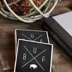 May include: Two black square coasters with white lettering and a white buffalo silhouette. The top coaster says "U F" and the bottom coaster says "B U F".