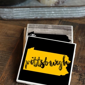 May include: A set of four black and white ceramic coasters with a yellow outline of the state of Pennsylvania and the word "pittsburgh" written in black. The coasters are on a wooden surface.