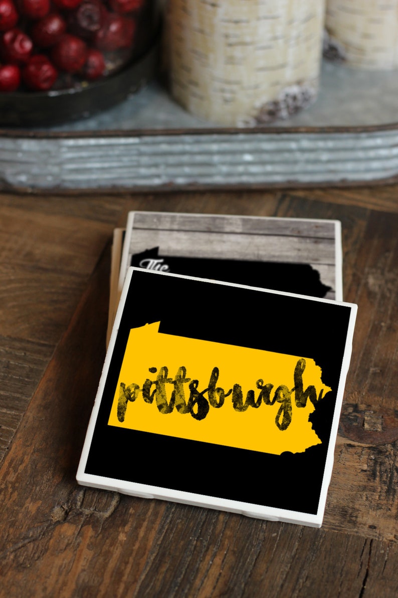 May include: A set of four black and white ceramic coasters with a yellow outline of the state of Pennsylvania and the word "pittsburgh" written in black. The coasters are on a wooden surface.