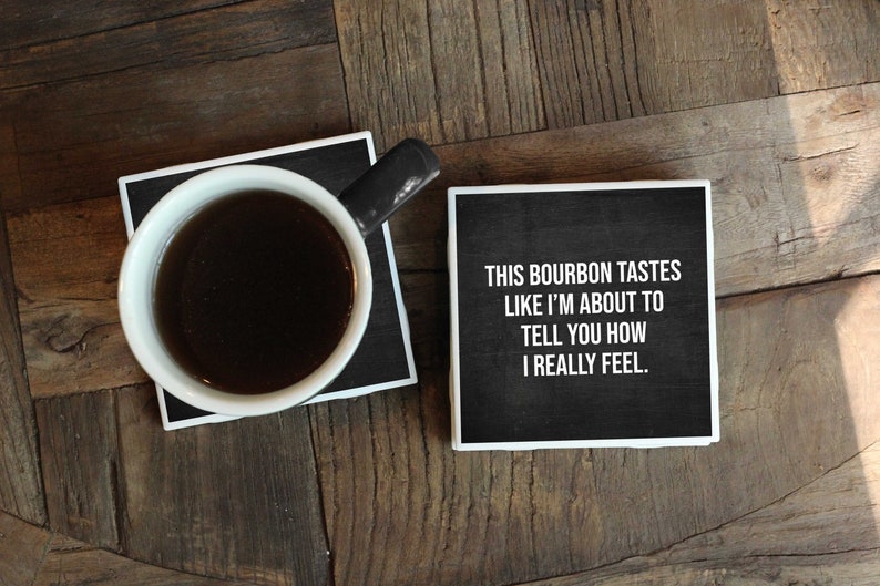 May include: Two black coasters with white borders. One coaster has a cup of coffee on it. The other coaster has the text "This bourbon tastes like I'm about to tell you how I really feel."