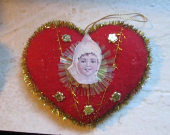 May include: A heart-shaped ornament with a red felt base, trimmed with gold tinsel. A vintage image of a child in a white bonnet is centered on the heart, surrounded by clear beads and gold accents. A gold string is attached for hanging.